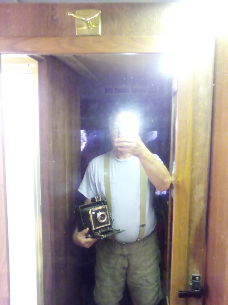 Man holding press camera under arm taking photograph in mirror with cell phone camera.